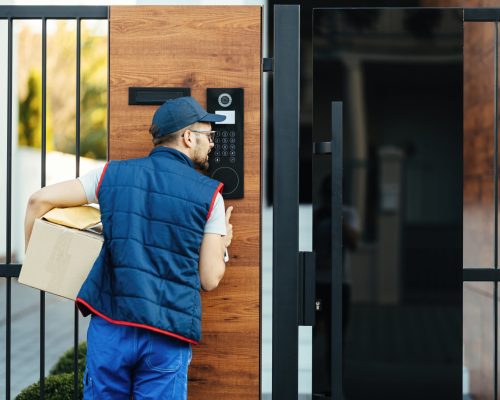 Back view of a deliverer ringing on intercom at gate of a customer's house while delivering packages.